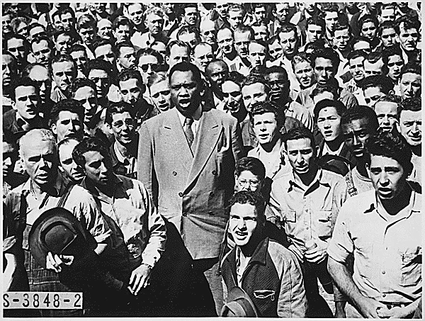 Paul Robeson standing among a crowd of shipyard workers as they sing together.